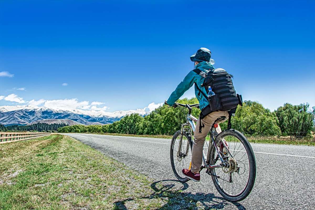 青空の下、舗装道路を自転車で走る男性の後ろ姿。自転車事故のリスクと予防意識を促すイメージ。