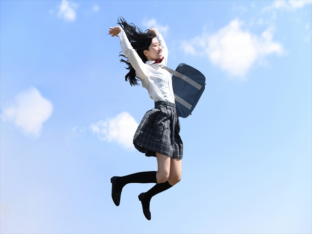 青空の下で両手を高く上げてジャンプする制服姿の女子高生
